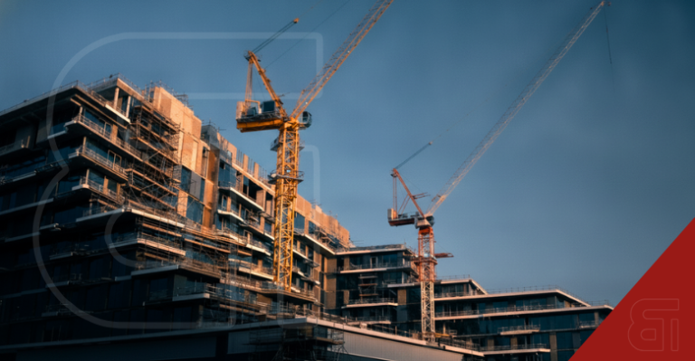 Two tall construction cranes operate above a large modern commercial building under development with scaffolding against a clear blue sky.